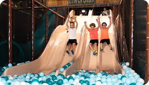 Three children racing down the side-by-side slides into the ball pit at Dinotopia