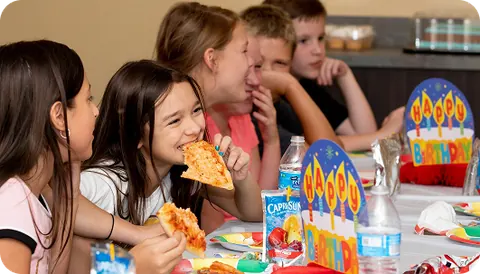 Children sitting around a table eating pizza together in the party room at Dinotopia