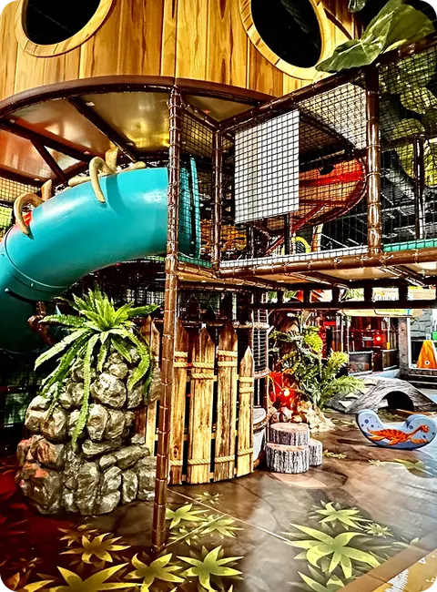 Inside Dinotopia: a turquoise tube slide winding out of a two-level themed climbing frame with rock and jungle detail