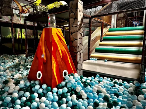 A deep ball pool filled with teal and white balls around a painted volcano, with soft padded steps leading out