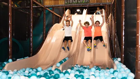 Three children racing down side-by-side wide slides into a teal and white ball pit