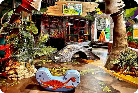 The toddler play area with a low dinosaur rocker, soft bridge and jungle planting under the Dinotopia Creek sign